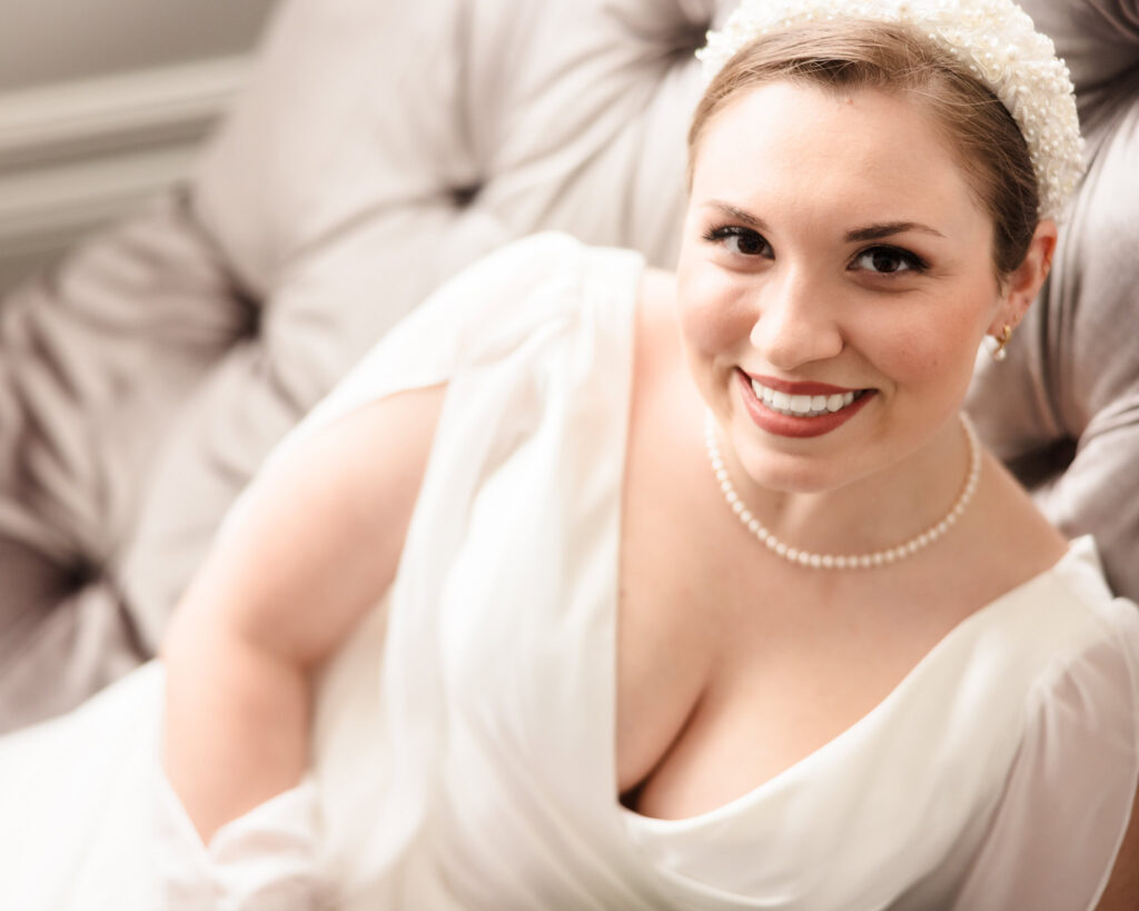 Close-up of a smiling bride in a pearl headband and white gown, seated on a velvet chair in the bridal suite.