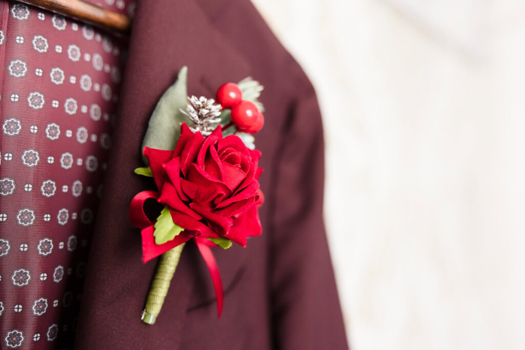Close-up of a burgundy groom’s suit with a red rose boutonniere featuring winter berries and pinecone accents for a wedding.
