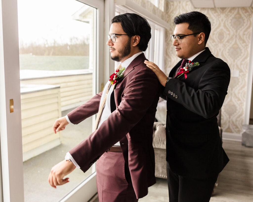 Groom in a burgundy suit getting ready with help from his groomsman in the groom’s suite at Giorgio’s Baiting Hollow before the winter wedding ceremony.