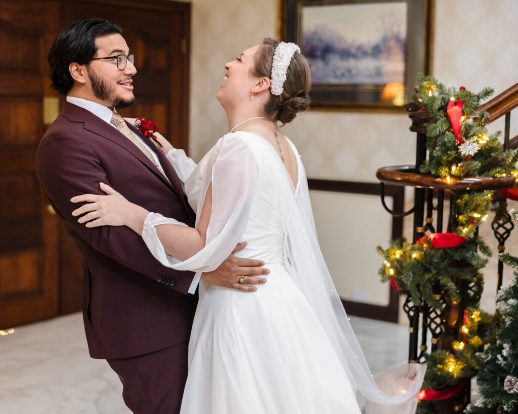 Bride and groom laughing and embracing during their first look beside the holiday-decorated staircase at Giorgio’s Baiting Hollow winter wedding.