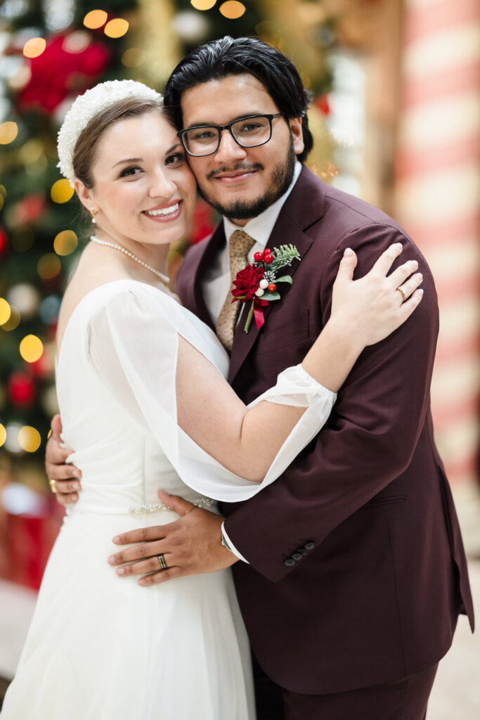 Bride and groom smiling and embracing in front of a Christmas tree at Giorgio’s Baiting Hollow, dressed in elegant winter wedding attire.
