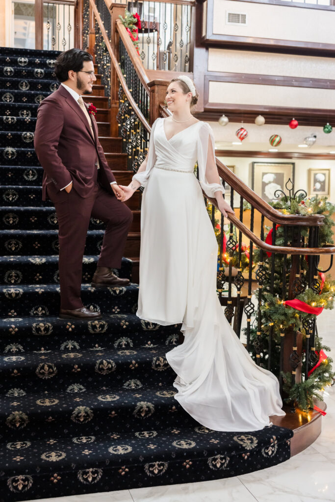 Bride and groom holding hands on the grand staircase at Giorgio’s Baiting Hollow, surrounded by festive holiday garland and warm winter decor.