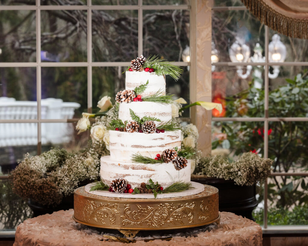 Rustic semi-naked winter wedding cake decorated with pinecones, greenery, and red berries on a gold stand at Giorgio’s Baiting Hollow reception.