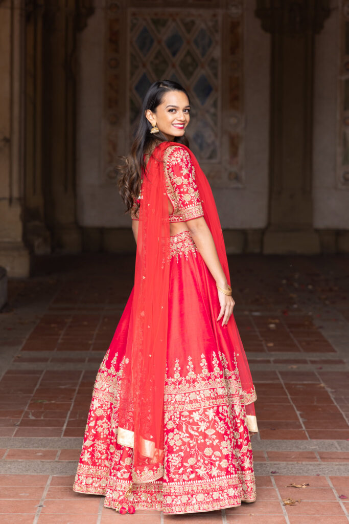 Woman smiling over her shoulder in traditional South Asian engagement attire, showcasing the detailed embroidery of her lehenga in Central Park.