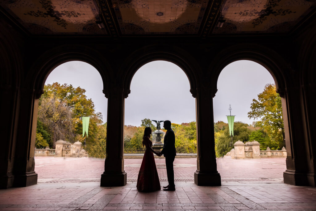 Silhouette of a couple holding hands in traditional South Asian engagement attire at Bethesda Terrace, with the Angel of the Waters fountain in the background.