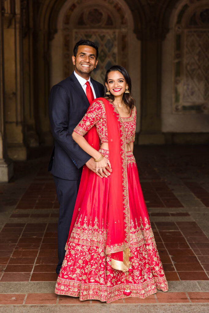 South Asian couple embracing at Bethesda Terrace during their Central Park engagement session, featuring a red lehenga and classic navy suit.