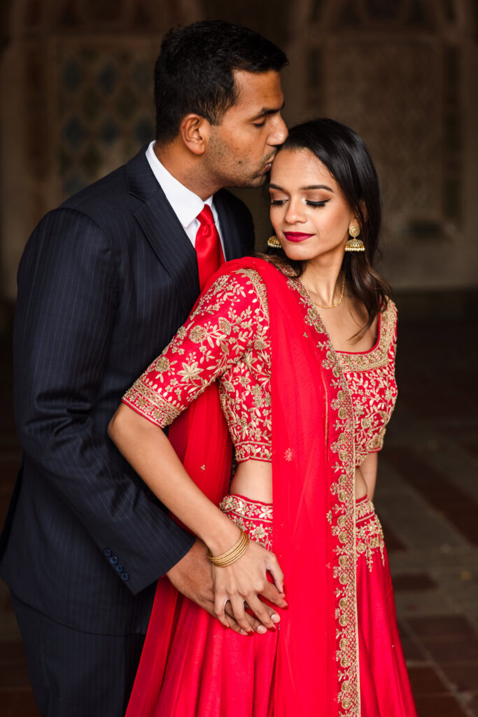 Groom-to-be kisses his fiancée’s forehead as they embrace in traditional Indian engagement attire at Bethesda Terrace.