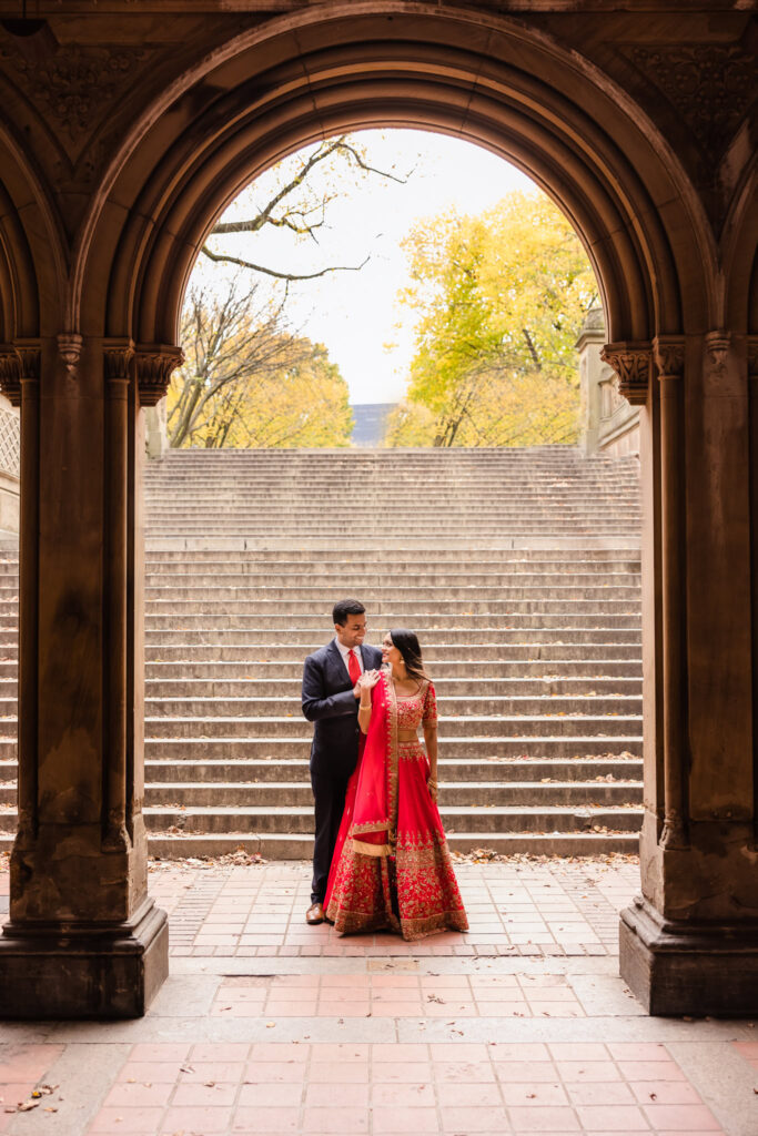 South Asian couple framed under the archway at Bethesda Terrace, with fall foliage and Central Park steps in the background during their engagement session.