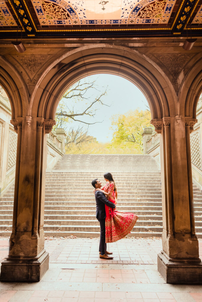 Groom-to-be lifts his fiancée beneath the ornate arches of Bethesda Terrace during their Central Park engagement session in vibrant South Asian attire.