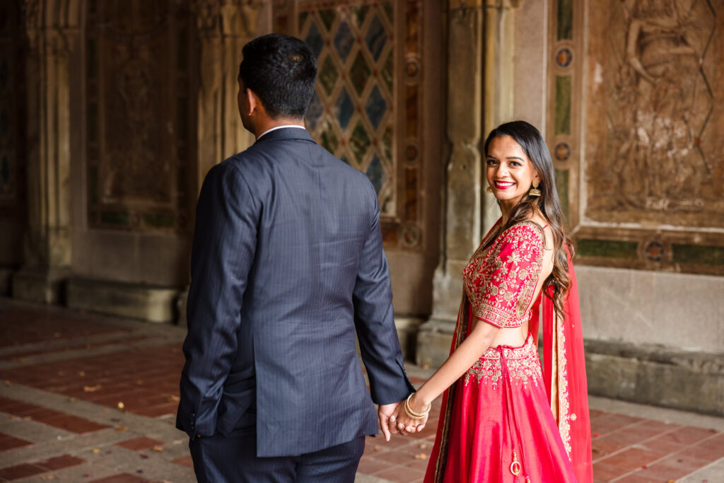 Bride-to-be in traditional South Asian attire smiles while holding hands with her fiancé at Bethesda Terrace during their cultural engagement session in Central Park.
