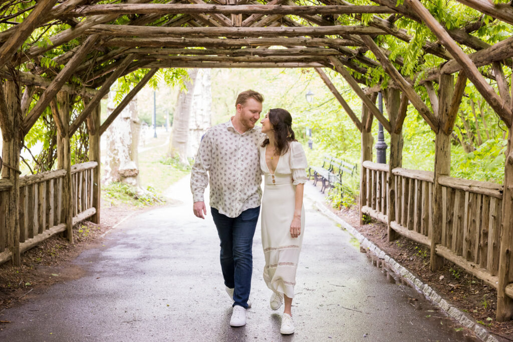 Playful couple walking under a rustic wooden arbor in Central Park, sharing a candid and joyful moment during their spring engagement session.