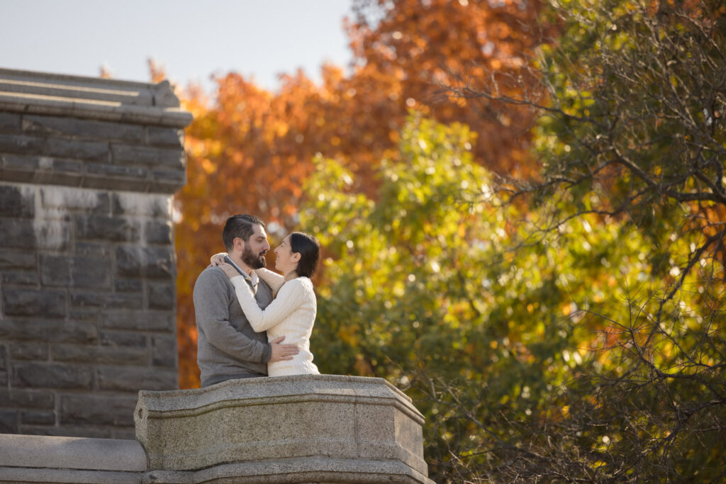 Engaged couple embraces on a stone balcony at Belvedere Castle in Central Park, surrounded by vibrant autumn foliage and warm afternoon light.