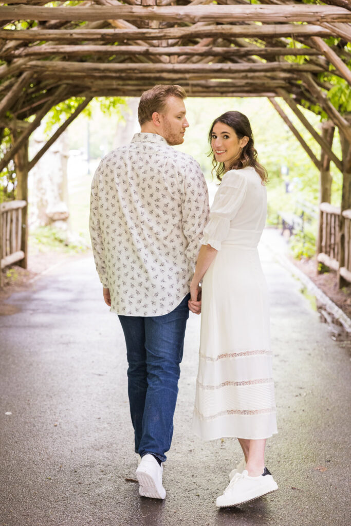 Couple holding hands and walking under a vine-covered wooden arbor in Central Park, with the bride-to-be smiling back during their spring engagement session.