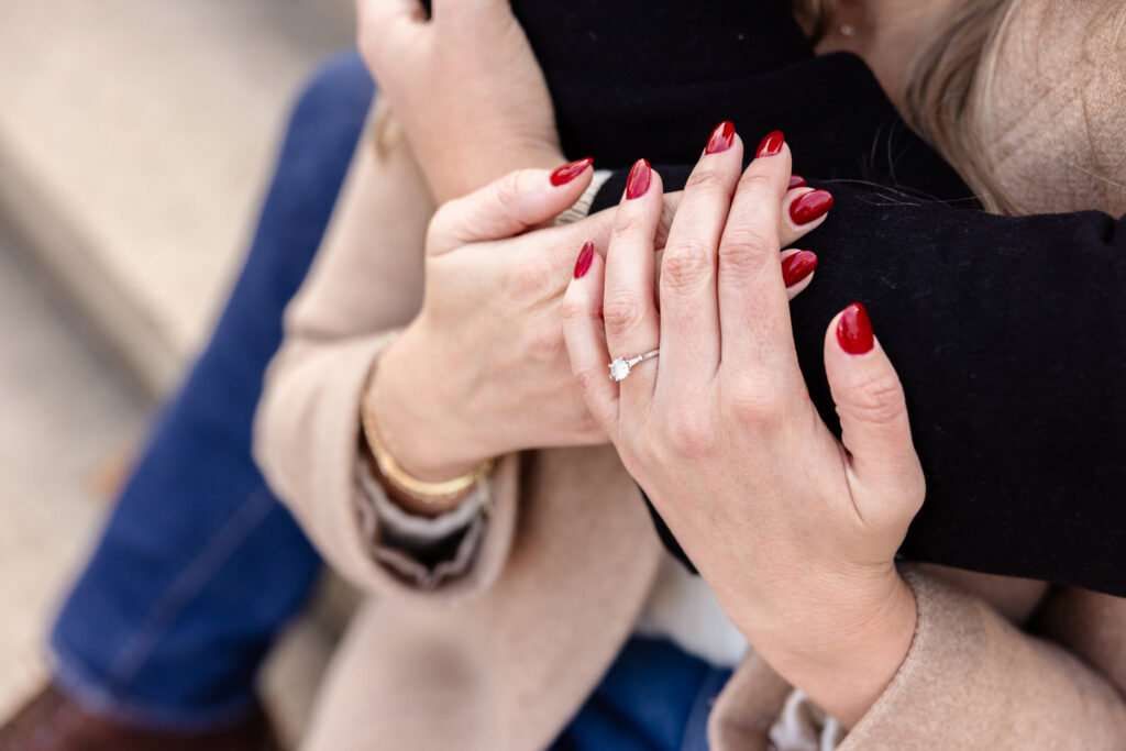 Close-up of bride-to-be’s hand with red nails and solitaire engagement ring resting on her partner’s arm during a winter Central Park engagement session.
