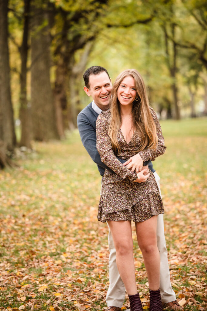 Laughing couple embraces in a playful moment among autumn leaves during their engagement session in Central Park’s tree-lined Mall.