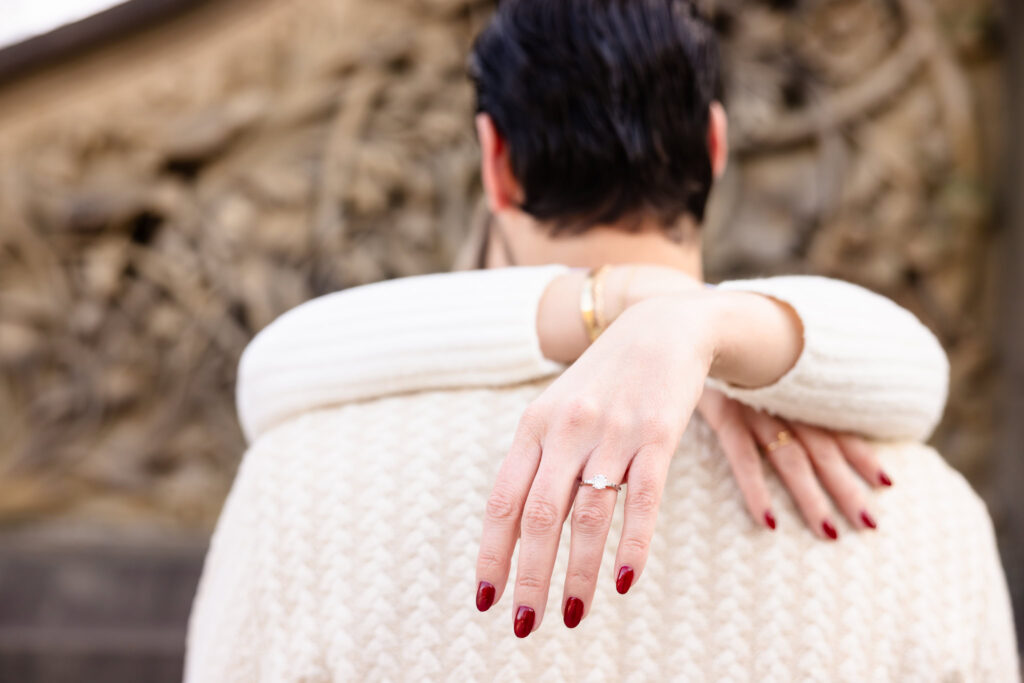 Close-up of bride-to-be’s red-manicured hand showing off her diamond engagement ring as she embraces her partner during a winter session at Bethesda Terrace.
