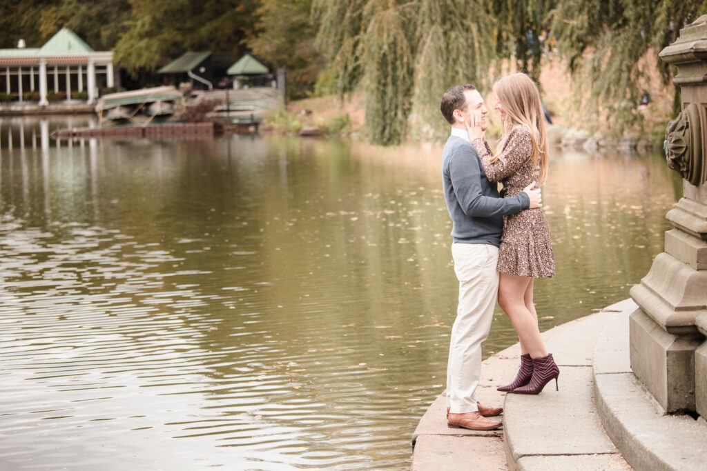 Joyful couple embraces by the edge of the Central Park Lake near Bethesda Fountain, sharing a playful moment during their autumn engagement session.