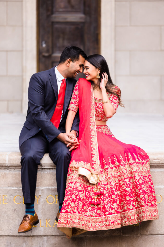 Couple sits on a stone ledge in Central Park, smiling warmly at each other as the bride-to-be wears a vibrant red and gold lehenga and the groom-to-be matches in a red tie.
