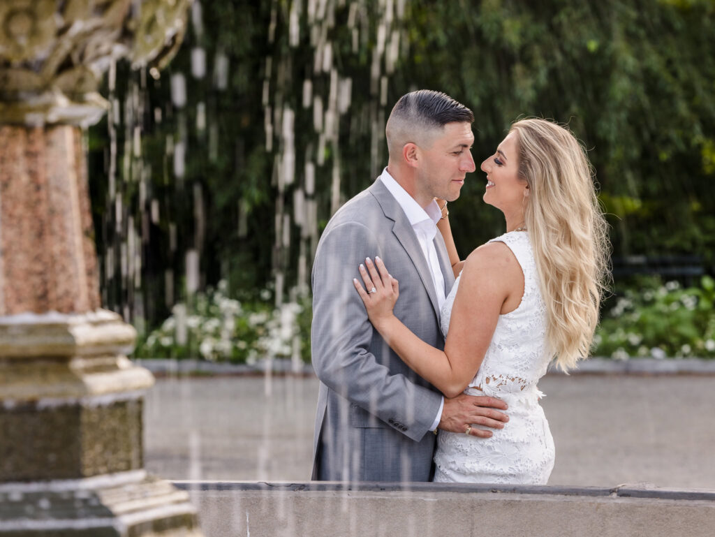 Engaged couple shares a romantic gaze beside the Conservatory Garden fountain in Central Park, framed by soft streams of falling water.