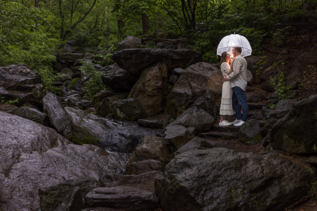 Couple shares a romantic moment under a glowing umbrella amid the rain-soaked rocks and lush greenery of Central Park's The Ravine during their moody engagement session.