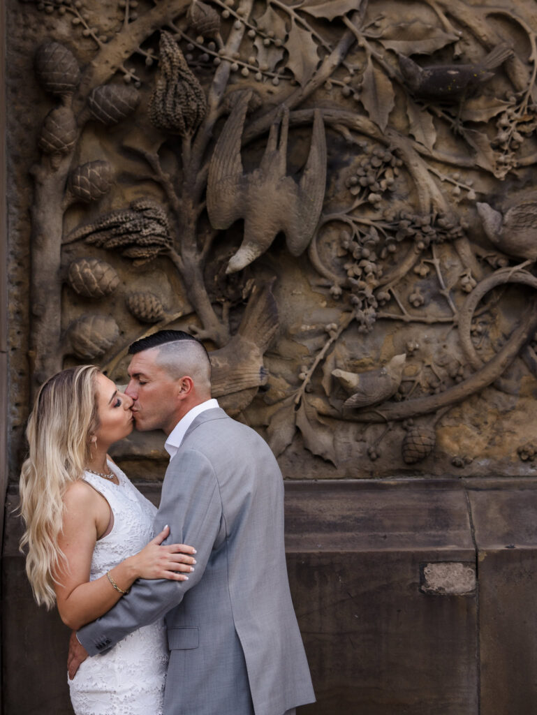 Couple shares a kiss in front of the intricate bird and foliage relief at Bethesda Terrace, highlighting their engagement with elegant outfits and romantic energy.