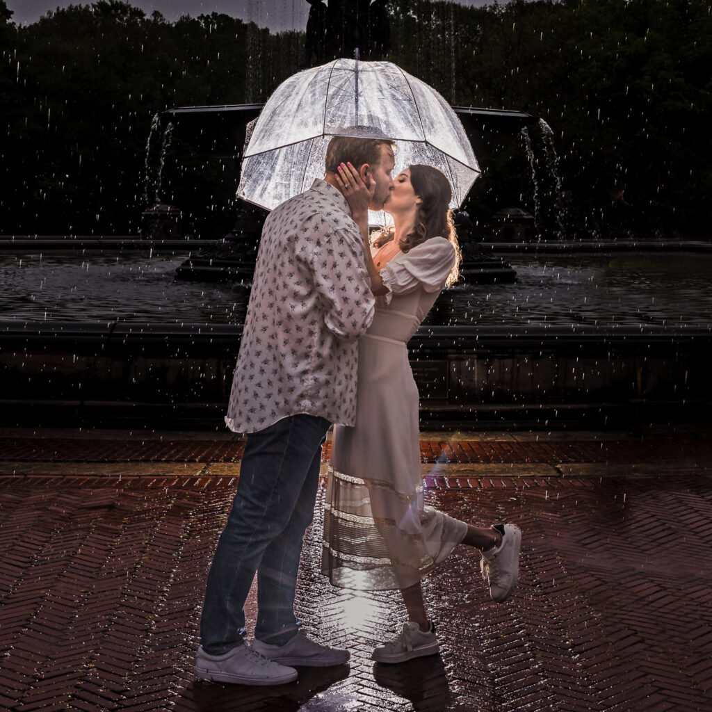 Couple kisses under a glowing umbrella in the rain at Bethesda Fountain, with dramatic backlighting capturing the romance of their rainy Central Park engagement session.
