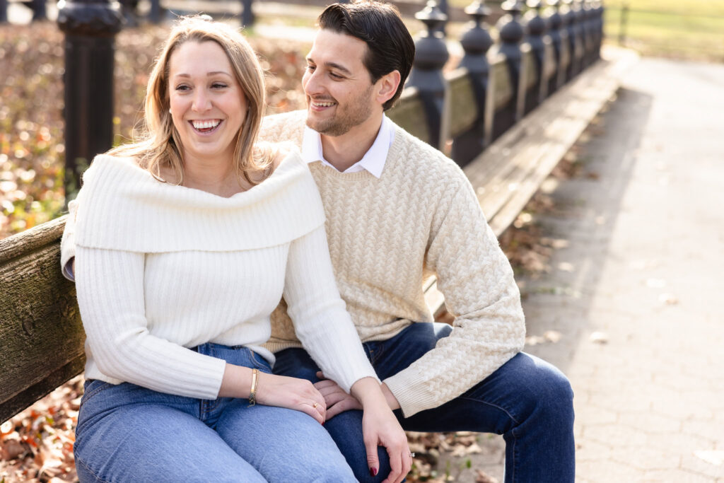 Engaged couple shares a cozy moment on a Central Park bench during winter, dressed in cream sweaters and jeans, smiling warmly in soft morning light.