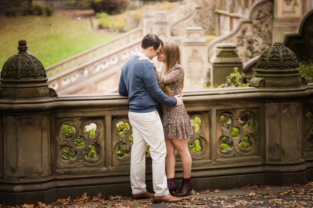 Romantic fall engagement photo of a couple embracing at Bethesda Terrace in Central Park, surrounded by ornate architecture and fallen leaves.