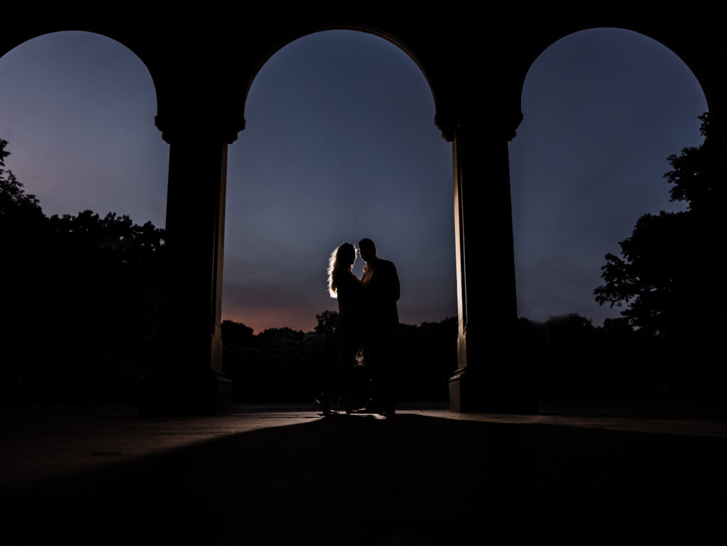 Silhouetted couple sharing an intimate moment beneath Bethesda Terrace arches during sunset in Central Park, capturing a dramatic and timeless engagement portrait.