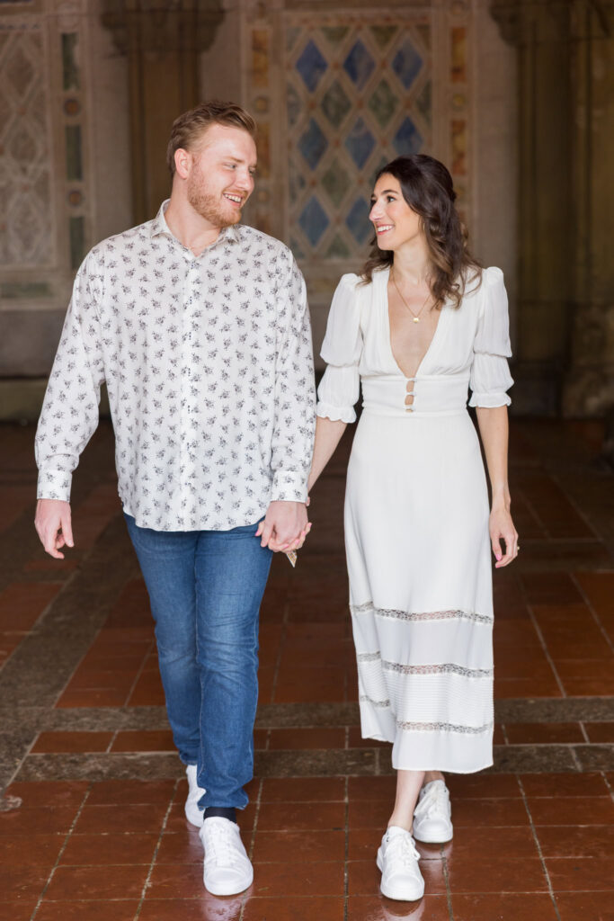Engaged couple holding hands and walking through Bethesda Terrace, smiling at each other during a relaxed spring engagement session in Central Park.