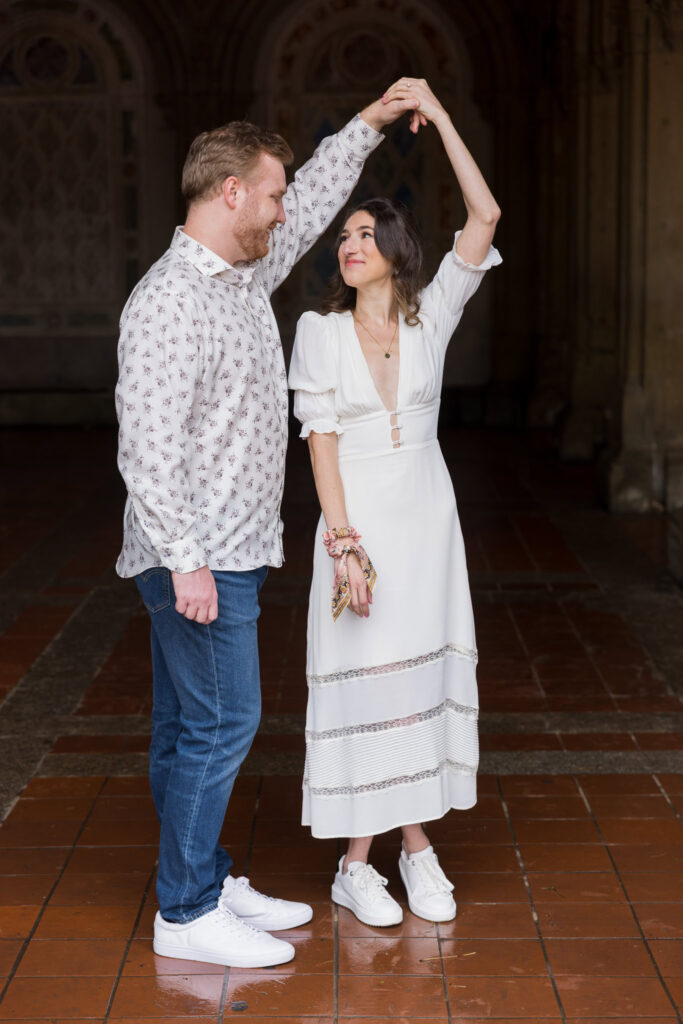 Groom-to-be twirling his fiancée under the arches of Bethesda Terrace during a playful moment in their rainy spring Central Park engagement session.