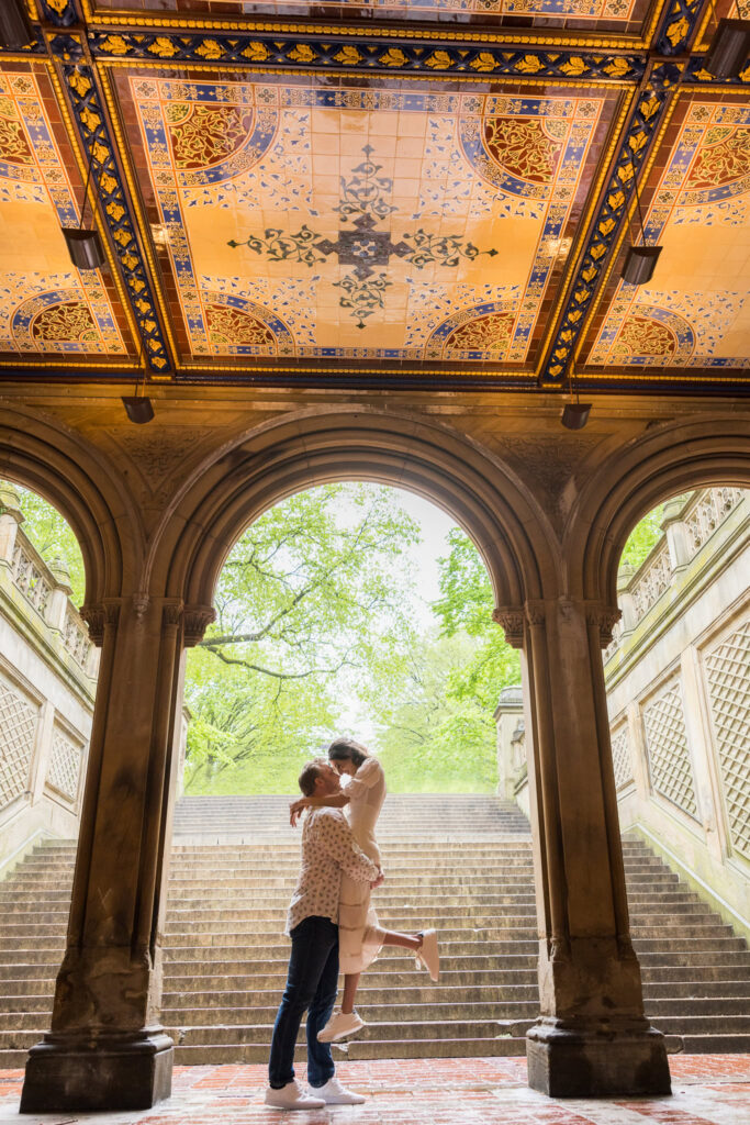 Groom lifting his bride-to-be for a kiss beneath the iconic Minton tile ceiling at Bethesda Terrace during their spring engagement session in rainy Central Park.