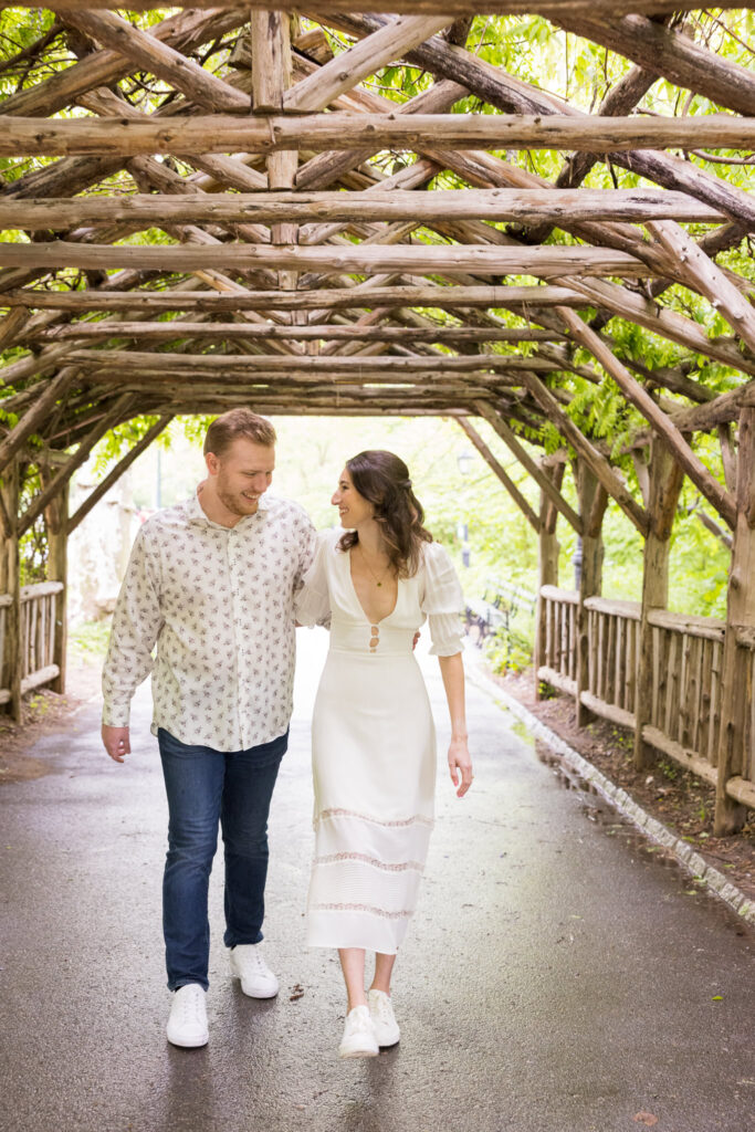 Engaged couple walking closely under a vine-covered wooden pergola in Central Park, sharing a joyful moment during their spring engagement session.