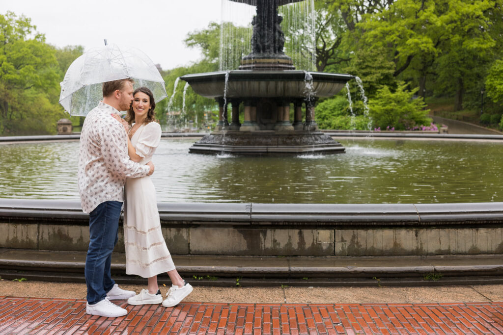 Engaged couple embracing under a clear umbrella in front of Bethesda Fountain during their spring engagement session in rainy Central Park.