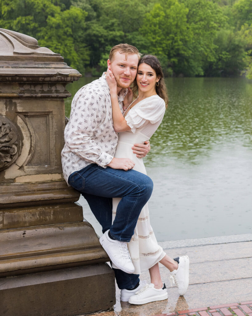 Couple embracing by the edge of Bethesda Terrace with raindrops falling on the lake during their spring engagement session in Central Park.