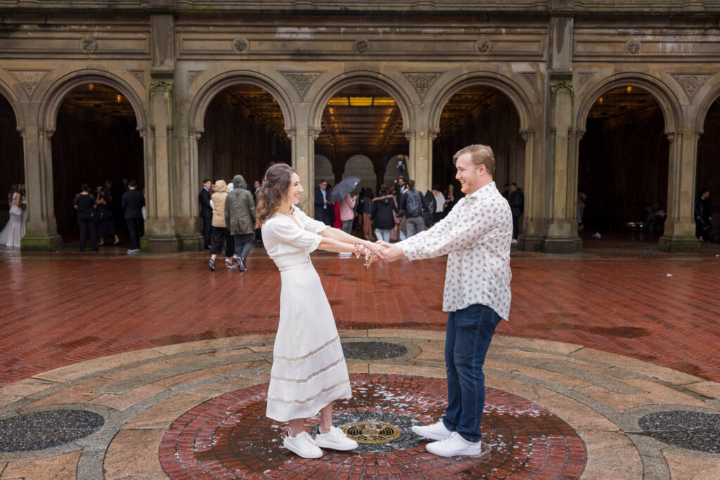 Couple laughing and holding hands in the center of Bethesda Terrace during a lively spring engagement session in the rain in Central Park.