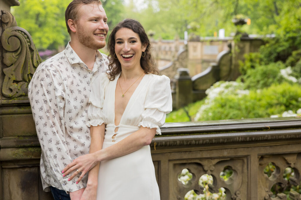 Couple laughing together on Bethesda Terrace overlooking the lush greenery of Central Park during their spring engagement session in the rain.