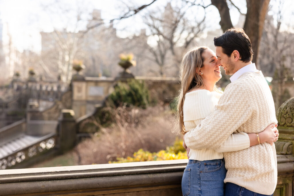 Engaged couple embracing and smiling at Bethesda Terrace in Central Park during a sunny winter engagement session.