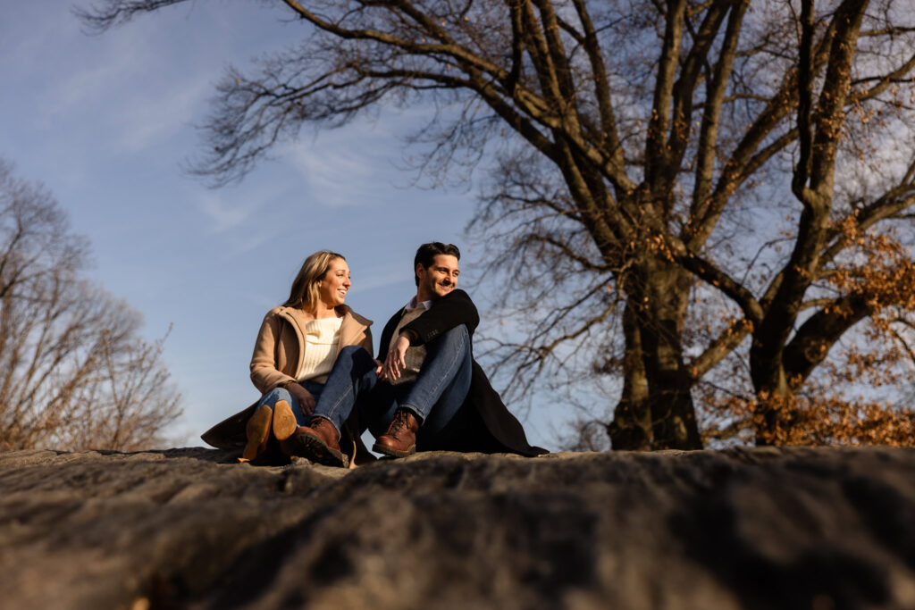 Couple sitting on a rock in Central Park during their winter engagement session, surrounded by bare trees and golden afternoon light.