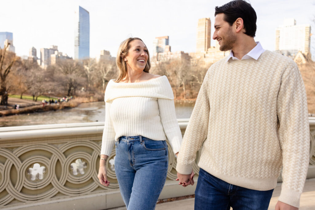 Couple holding hands and smiling at each other while walking across Bow Bridge during their winter engagement session in Central Park.