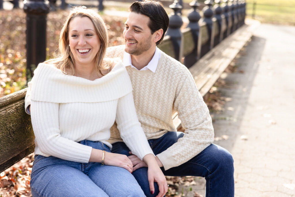 Couple sharing a joyful moment while sitting on a wooden bench in Central Park during their winter engagement session, dressed in cozy neutral sweaters.