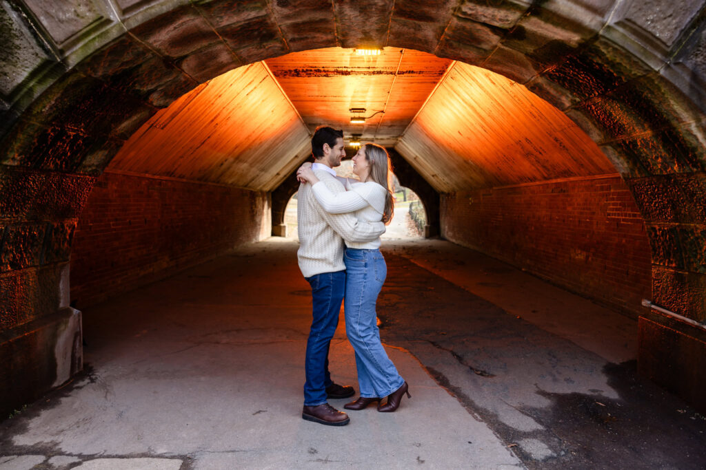 Couple embracing under a warmly lit stone and brick archway in Central Park during their winter engagement session.