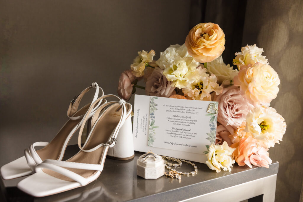 Flat lay of bridal details for a wedding, featuring white block heels, gold jewelry, floral wedding invitation, and a soft pastel bouquet perfect inspiration for a Long Island fall waterfront wedding.