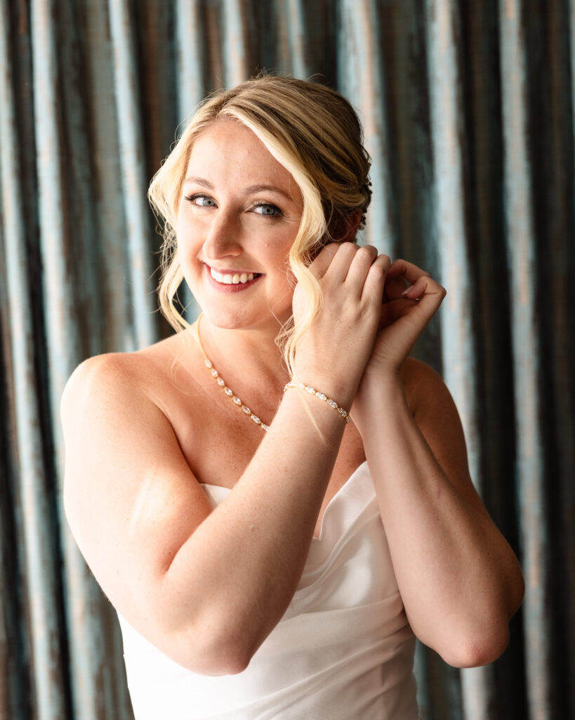 Bride smiles softly while putting on earrings before her wedding, radiating joy and elegance ahead of her Long Island fall waterfront ceremony.
