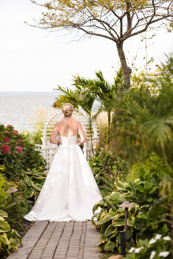 Bride walks through the lush garden path toward her first look at Crescent Beach Club, surrounded by greenery and waterfront views during her elegant September wedding on Long Island.