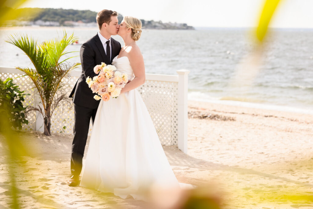 Bride and groom share a kiss on the sand with pastel bouquet in hand, framed by the shoreline at their Crescent Beach Club waterfront wedding on a warm September day on Long Island.