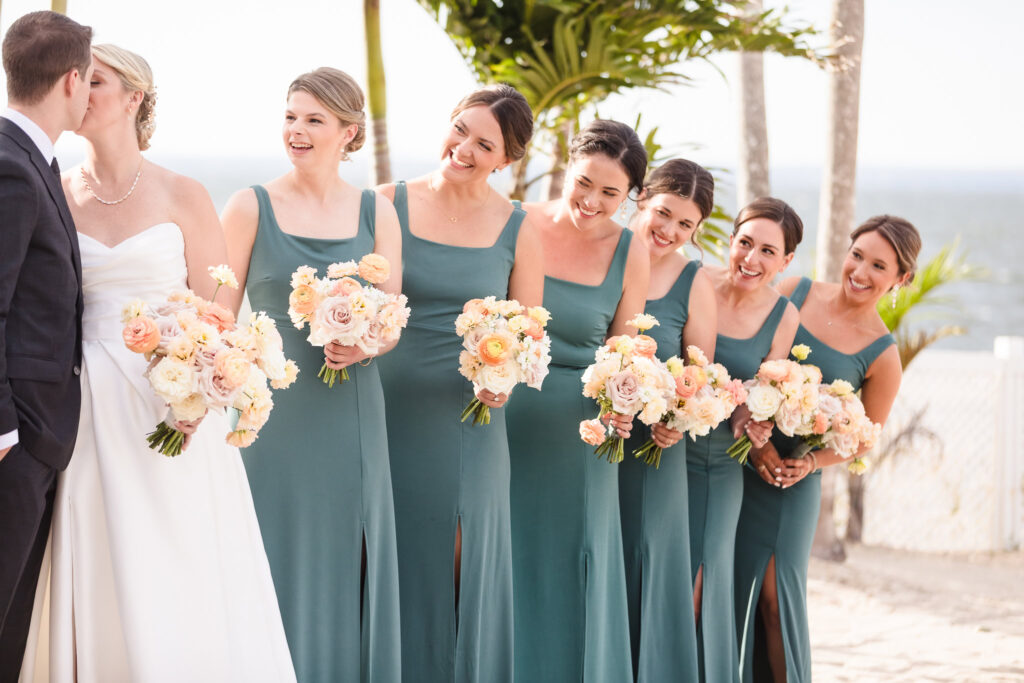 Bride and groom share a kiss as bridesmaids in sage green dresses smile beside them, holding pastel bouquets during a joyful Crescent Beach Club September wedding on the Long Island waterfront.