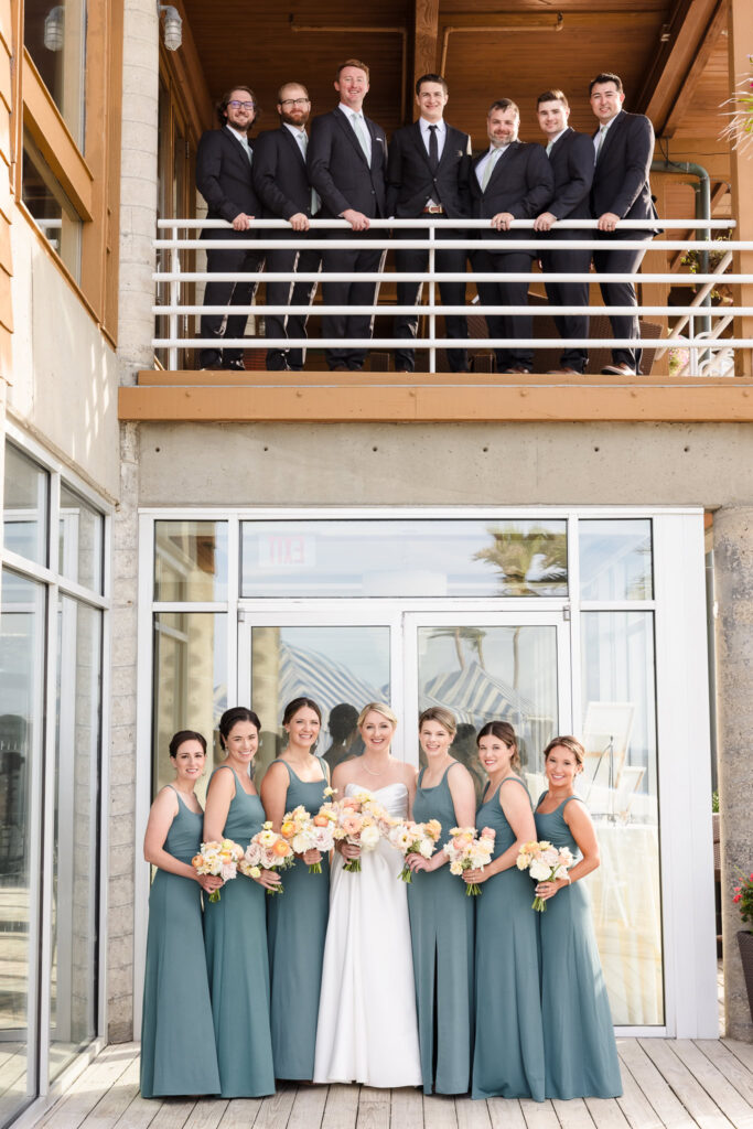 Wedding party portrait at Crescent Beach Club with groomsmen standing above on the balcony and bridesmaids in sea-glass gowns below, capturing a joyful September wedding moment on Long Island.