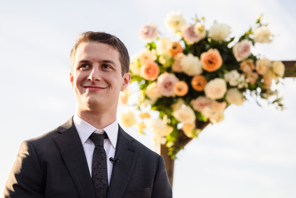 Groom smiles with anticipation beneath a floral arch at his ceremony, moments before his September wedding begins on the Long Island shore.
