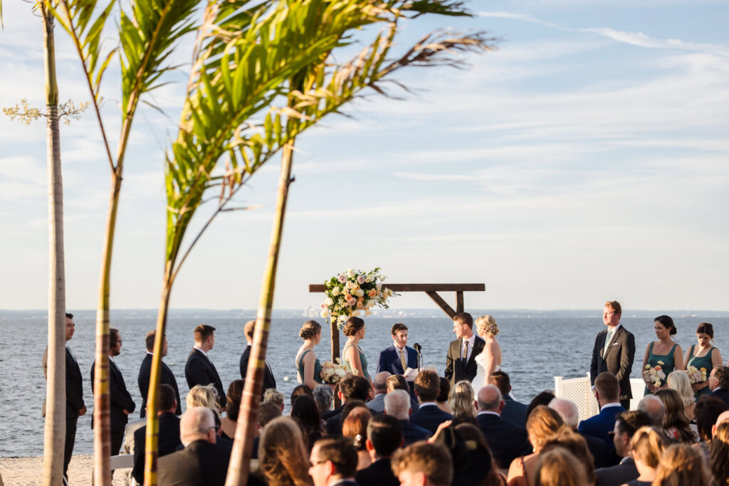 Crescent Beach Club waterfront ceremony with bride, groom, and wedding party under a floral arch, surrounded by guests and framed by palm trees on a serene September afternoon in Long Island.